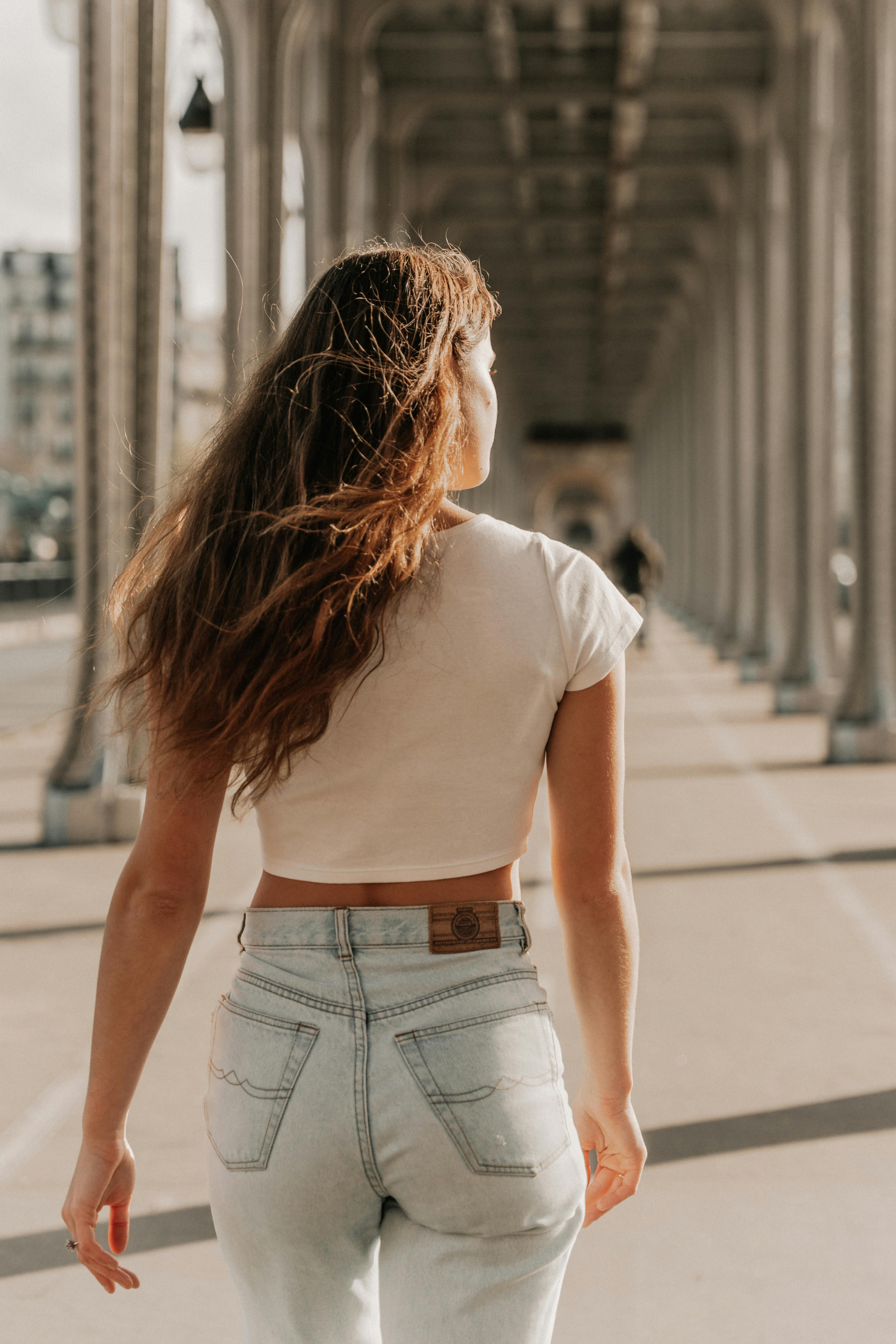 A stylish young woman with long brown hair walking under an urban bridge in daylight.