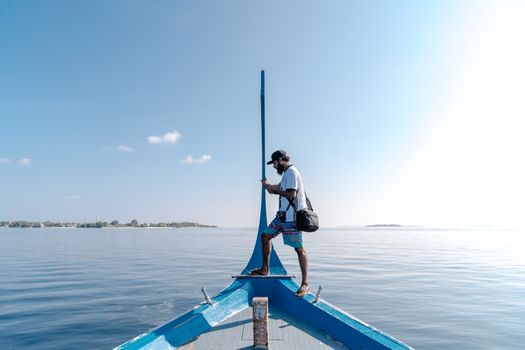 A man stands on a boat prow using a smartphone with serene sea and clear sky in the backdrop.