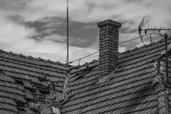 A grayscale photo of a broken roof with a chimney and antenna under a cloudy sky.