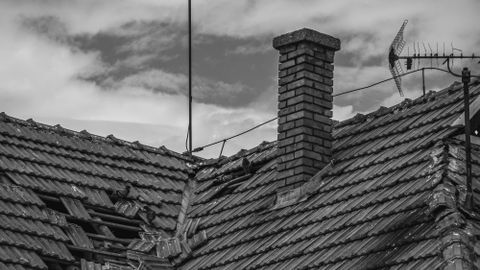 A grayscale photo of a broken roof with a chimney and antenna under a cloudy sky.