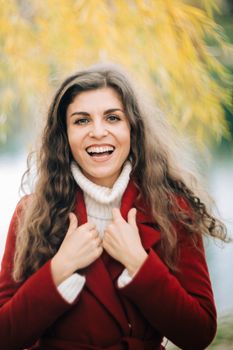 Happy woman with curly hair smiling in a red coat, enjoying a bright day outdoors.