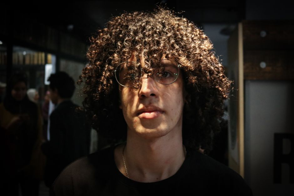 Close-up photo of a young man with curly hair and glasses, highlighting fashion style and expression.