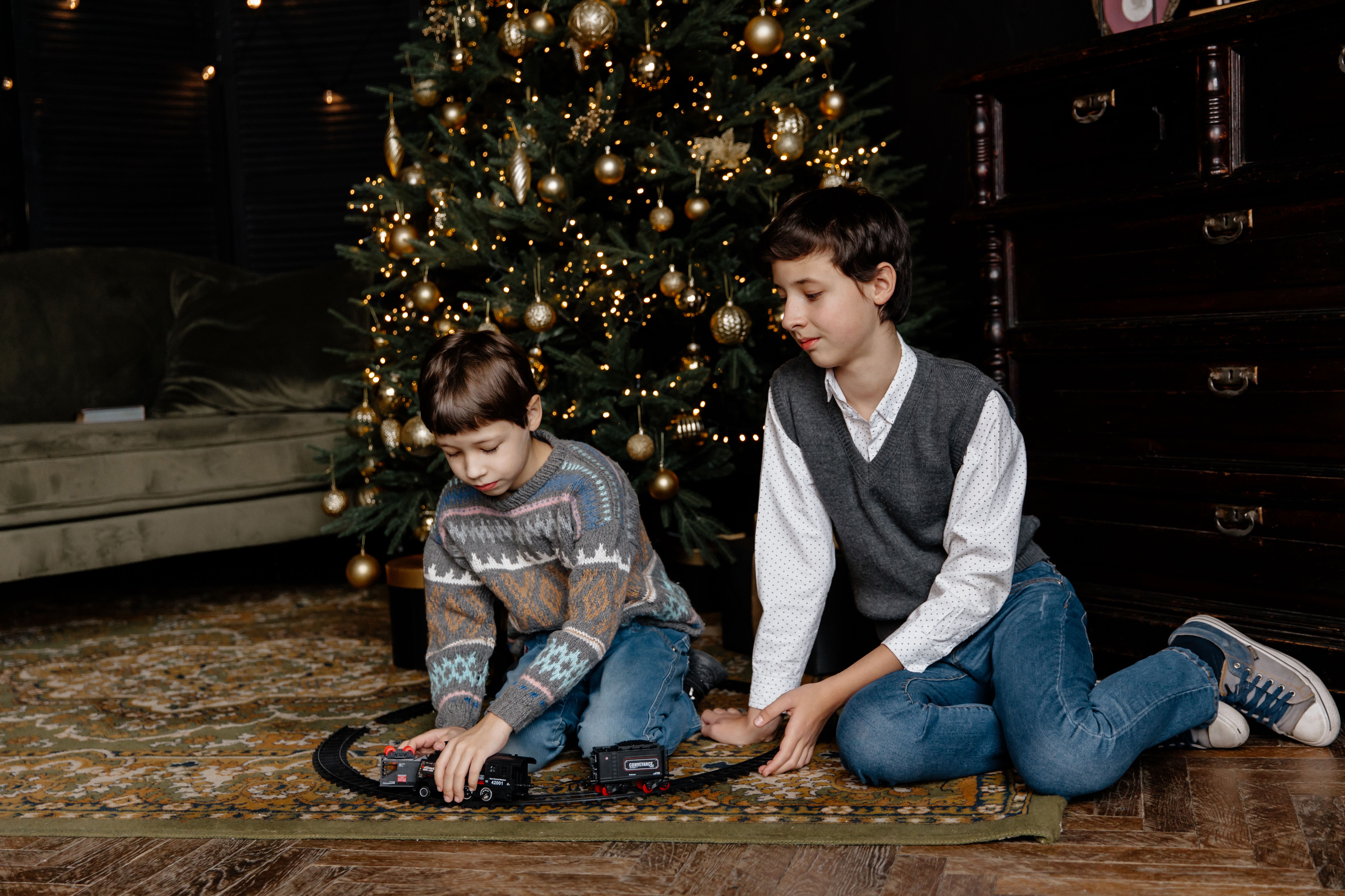 Two boys enjoying a playful moment with a toy train set near a beautifully decorated Christmas tree indoors.