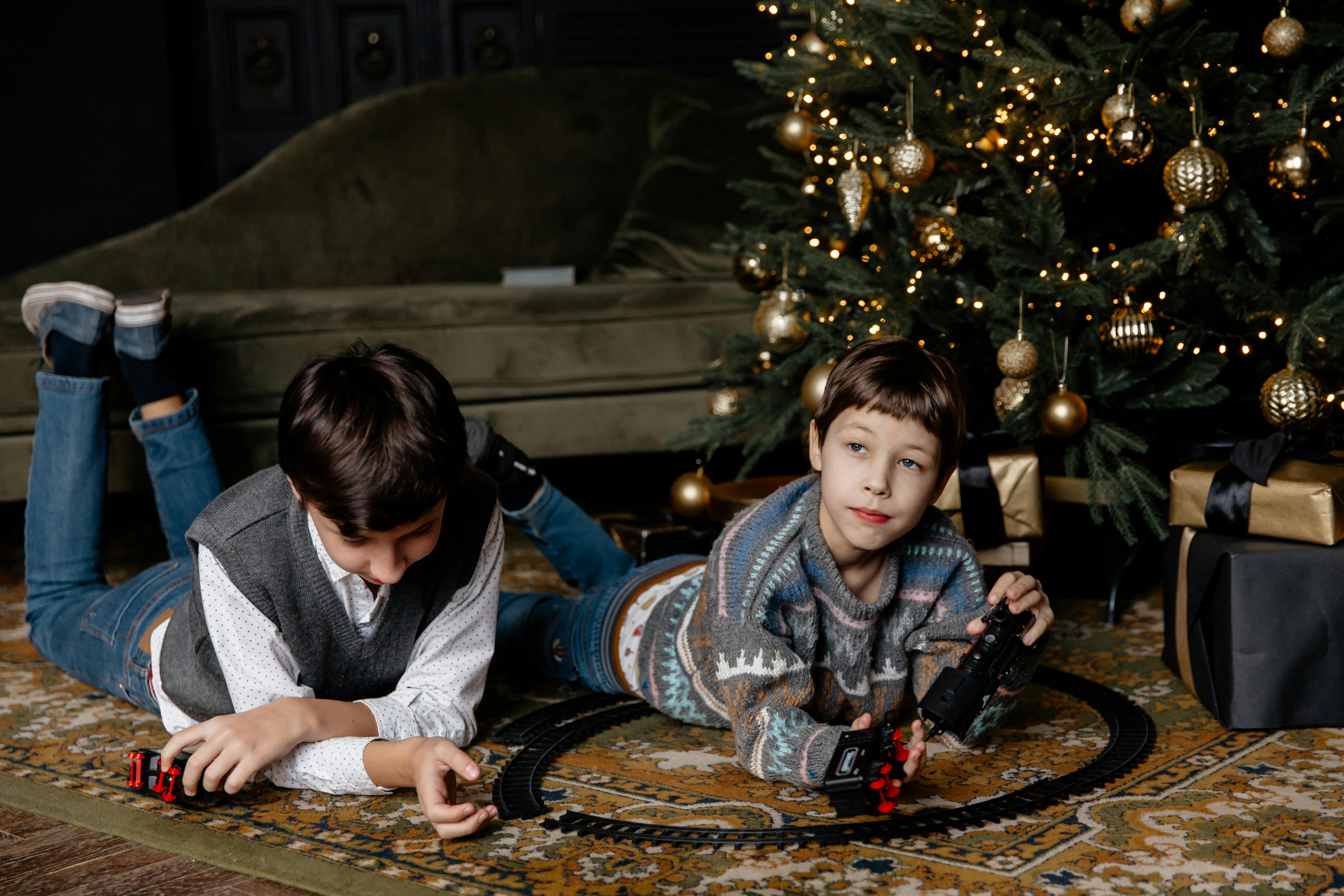 Two children enjoy playing with a toy train set next to a decorated Christmas tree indoors.