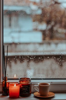 A cozy scene with coffee, candles, and rain-splattered window, perfect for relaxation.
