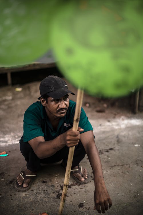 Free A Man in Green Shirt and Black Cap Holding a Wooden Stick while Sitting on the Street Stock Photo