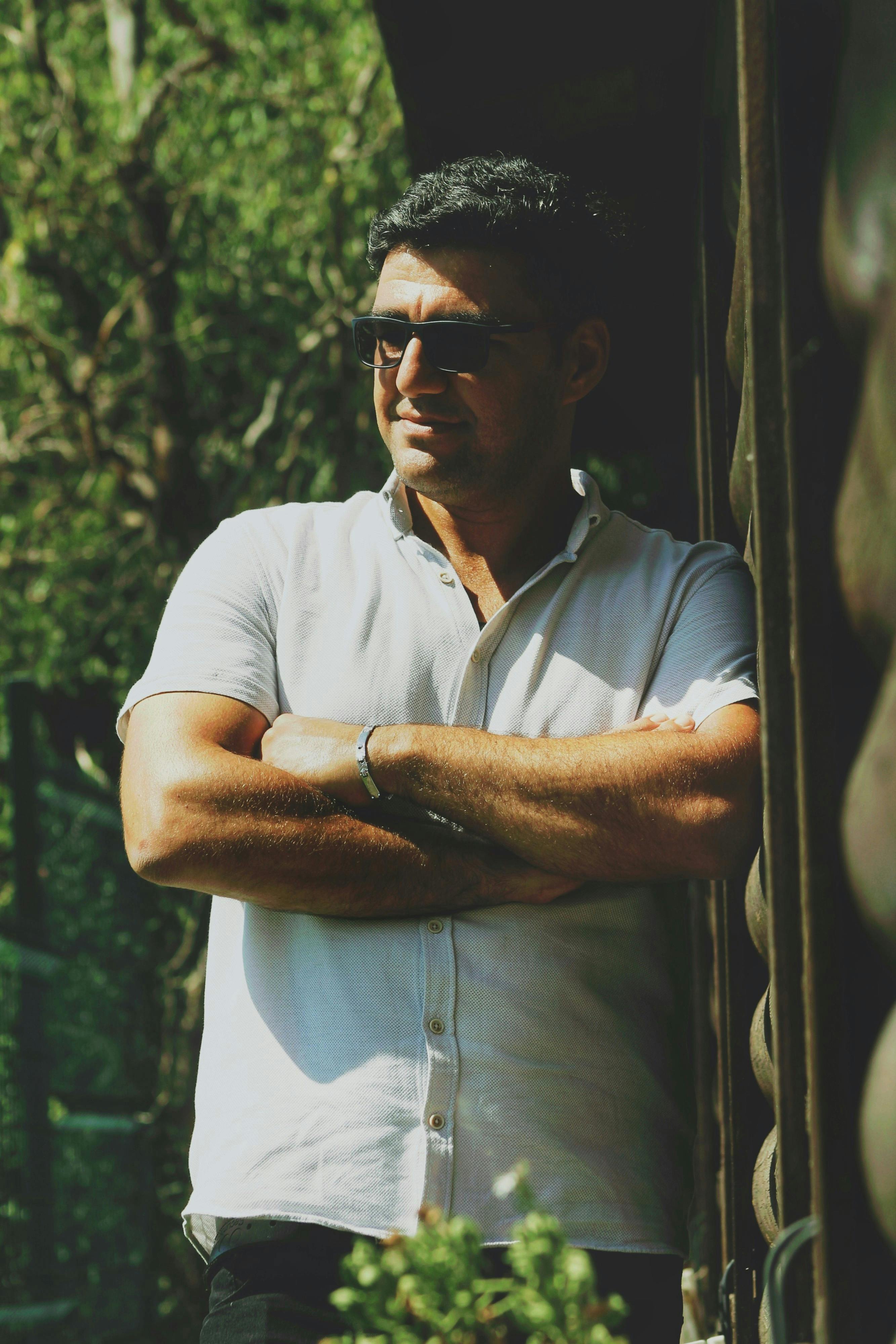 A man in sunglasses leans against a wall, arms crossed, under bright sunlight.