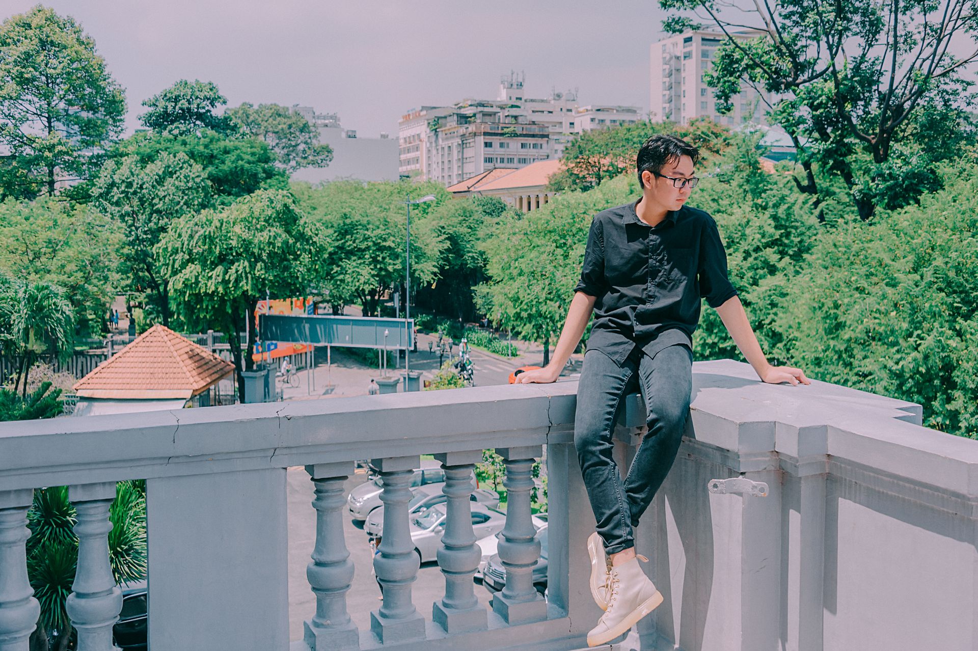 Casual man in black outfit sitting on a balcony amidst a lush urban landscape on a sunny day.