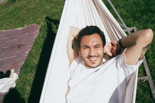 Free A relaxed, tattooed man smiles while lounging in a white hammock on a sunny day. Stock Photo