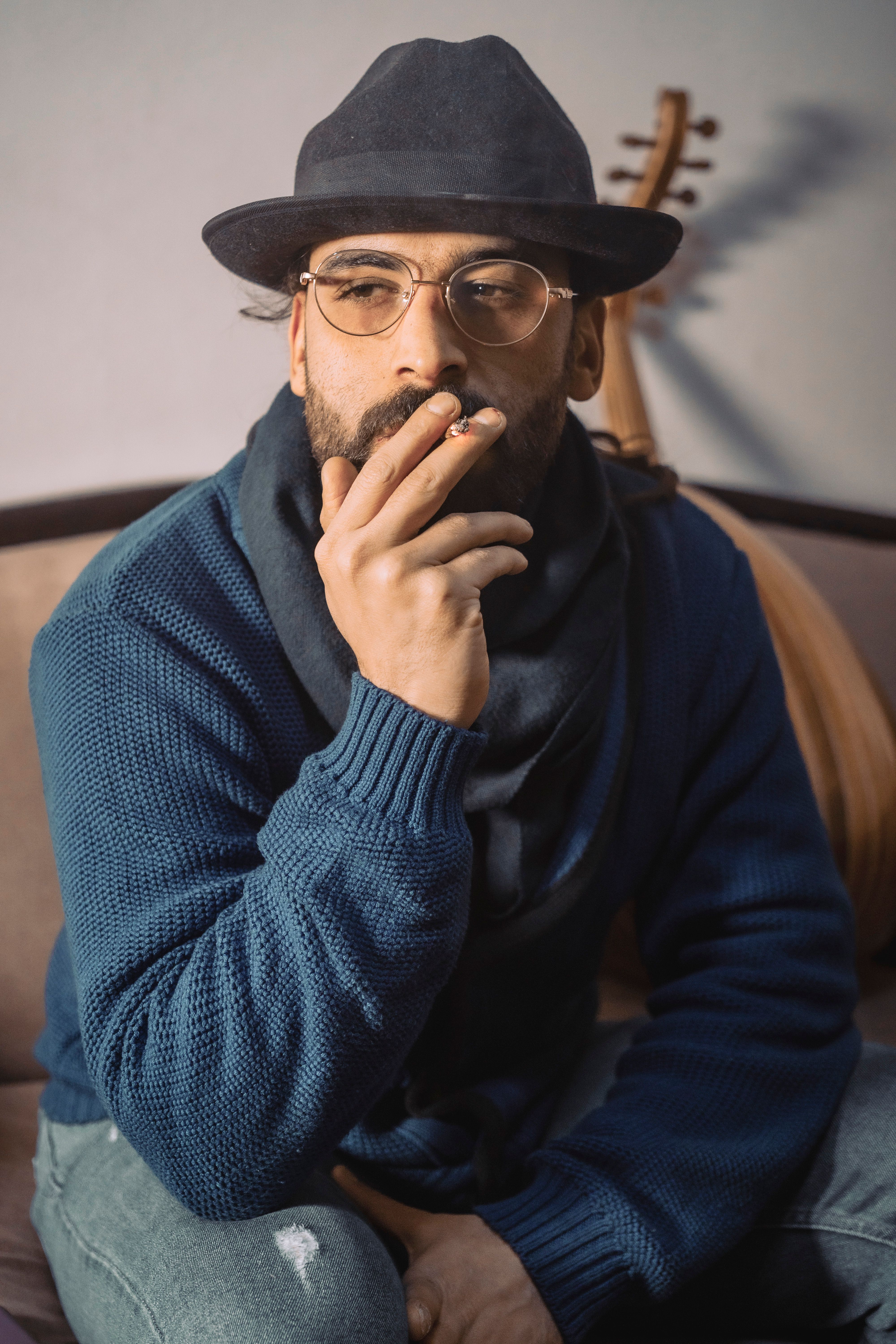 Bearded man in glasses wearing hat and blue cardigan smoking indoors with musical instrument.