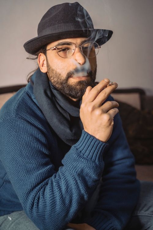 Free Portrait of a bearded man smoking indoors, wearing a hat, glasses, and a blue sweater. Stock Photo
