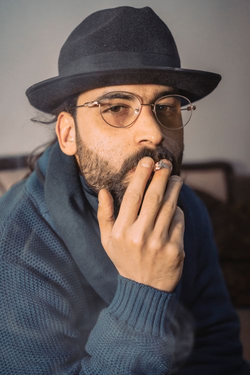Free Close-up of a bearded man wearing glasses and a hat, smoking indoors. Stock Photo