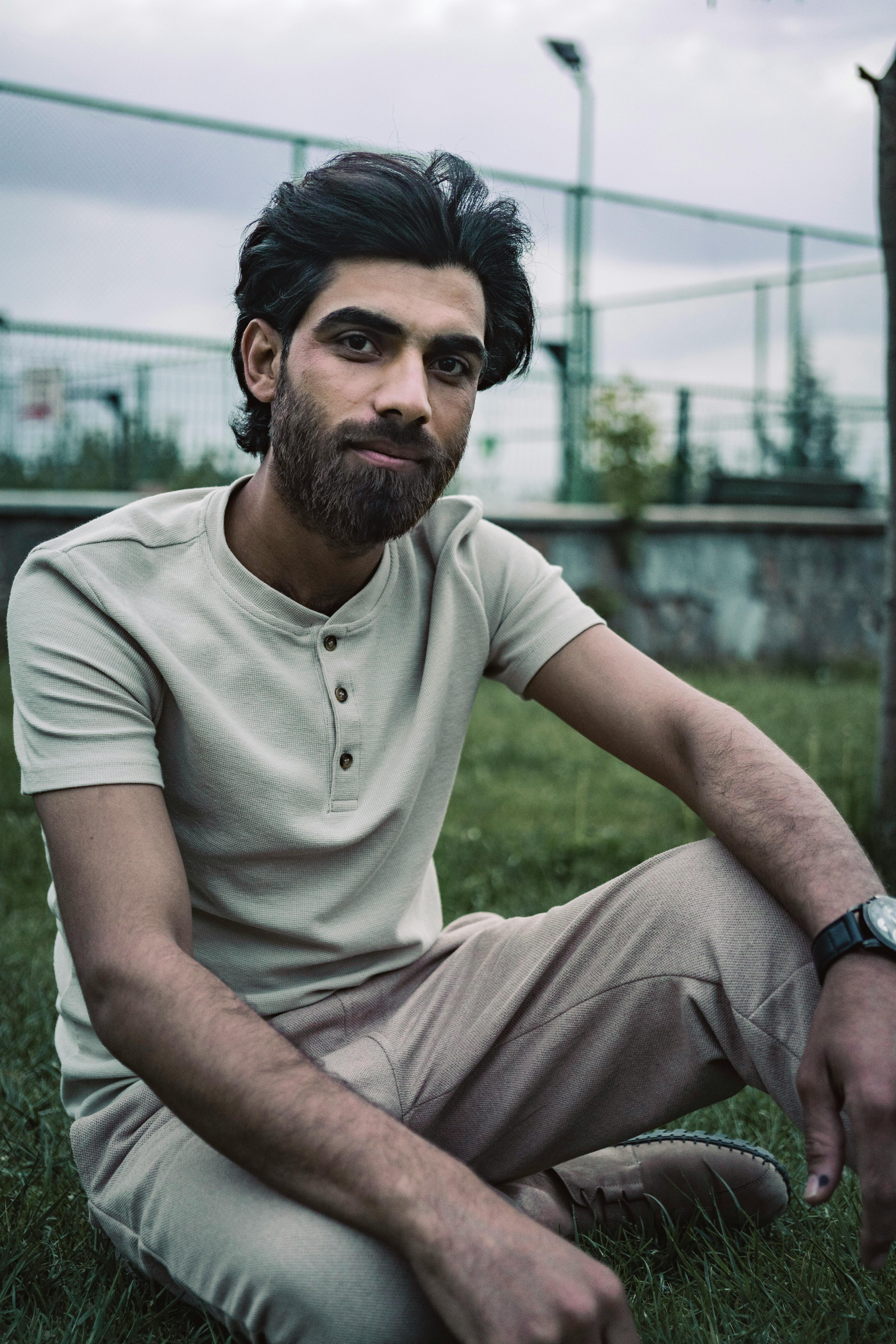 A relaxed portrait of a bearded man sitting on grass, showcasing urban style.