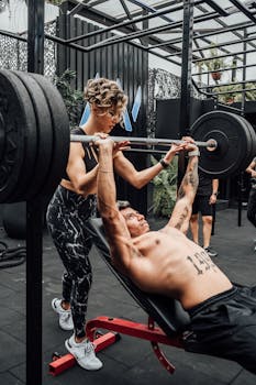 A muscular man performing a barbell bench press with assistance in a modern gym.