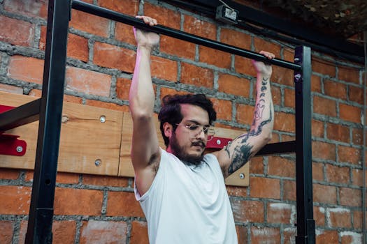 A man with tattoos and eyeglasses doing pull-ups inside a gym setting with a brick wall backdrop.