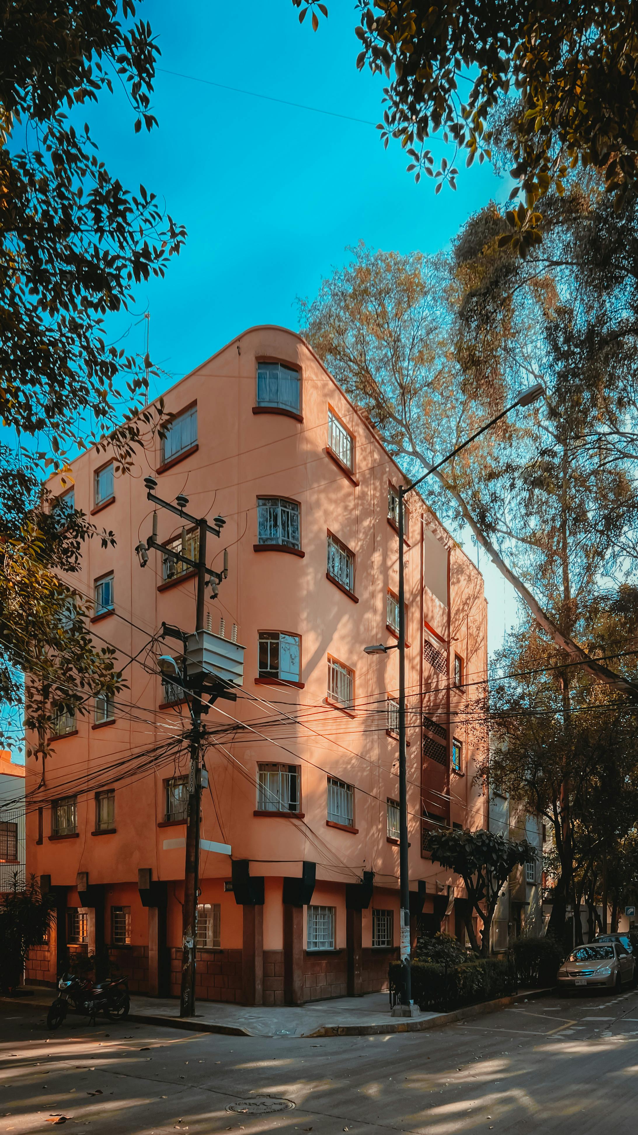 Free A vibrant pink apartment building on a street corner with lush green trees and clear blue sky. Stock Photo
