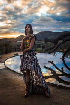 Fashionable woman in a patterned dress stands by a pool at sunset, exuding elegance and style.