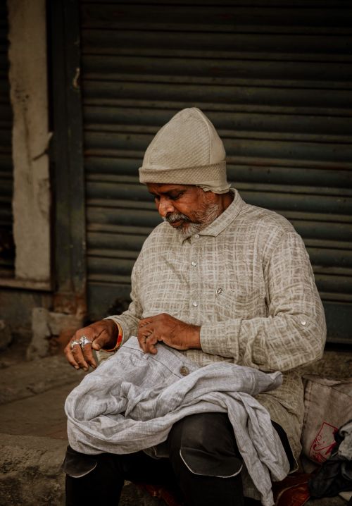 Free Elderly man with a plaid shirt and beanie, sitting on the street, sewing. Stock Photo