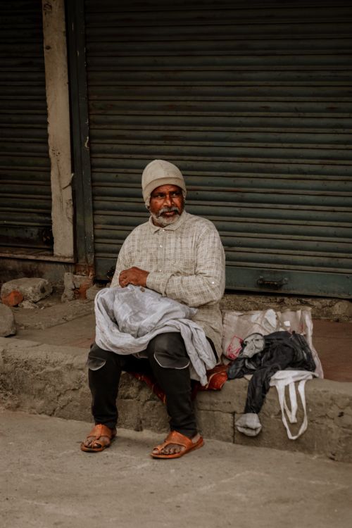 Free Elderly man seated on a Bengaluru street with clothes, shutters behind him, showcasing urban lifestyle. Stock Photo