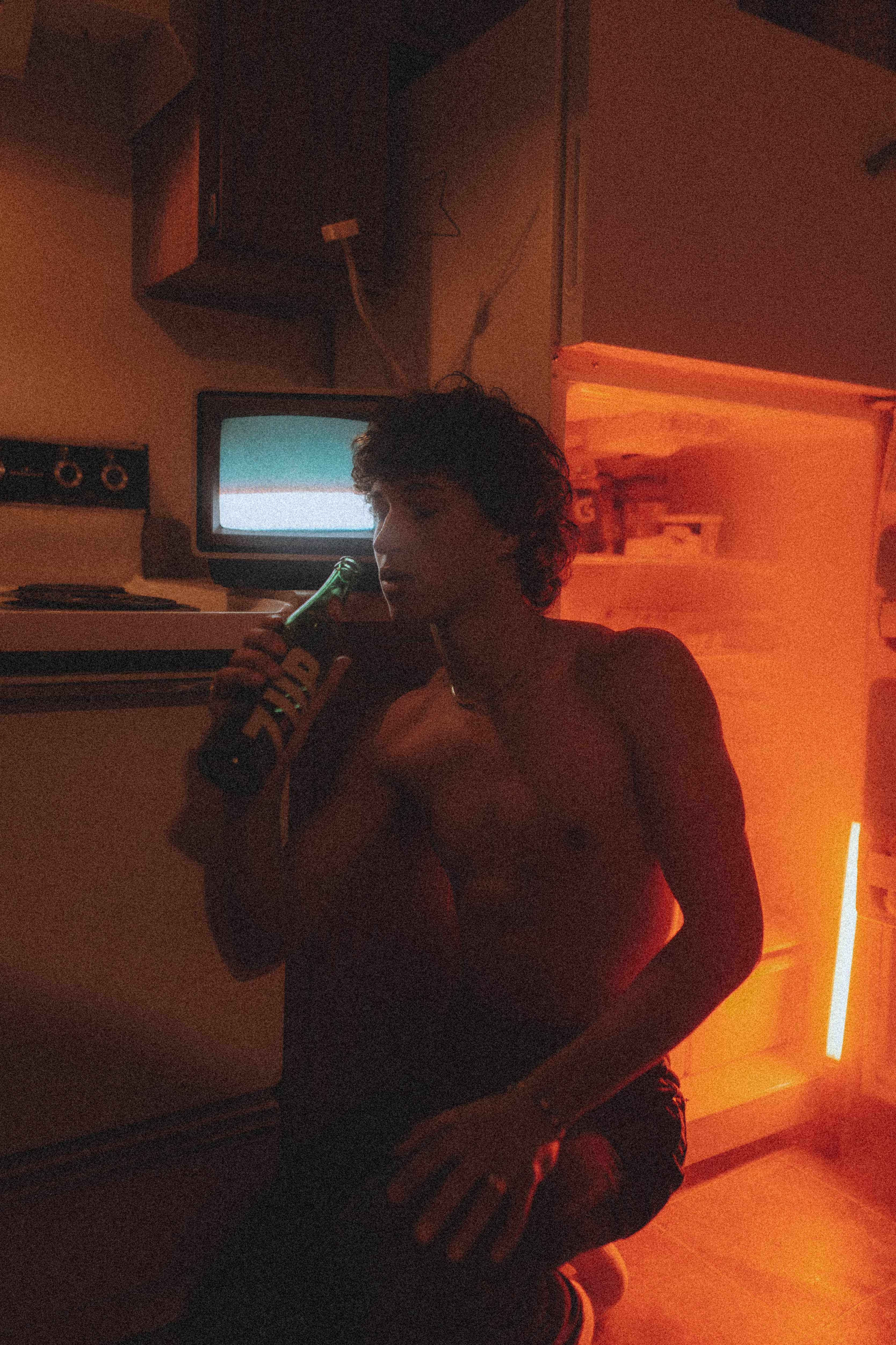 Shirtless man with curly hair enjoying a drink by the fridge with a moody, warm glow.