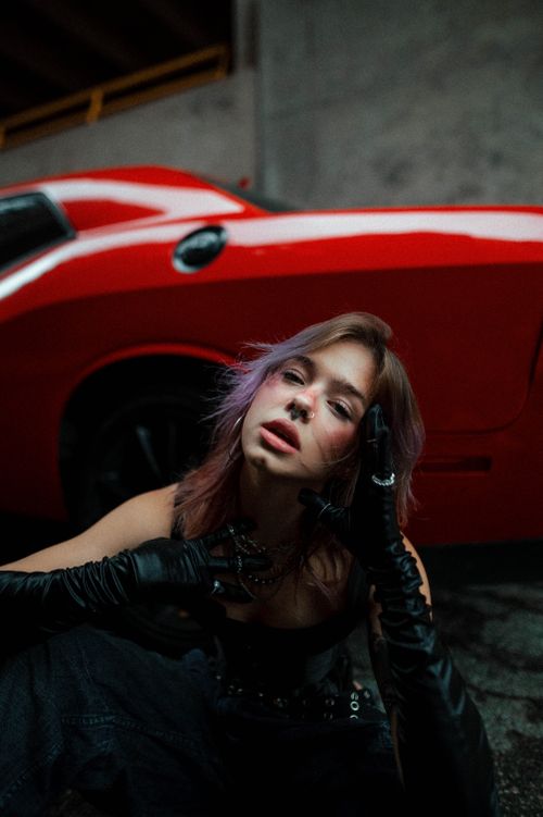 Free Stylish woman with purple hair poses by a striking red car in an urban setting. Stock Photo