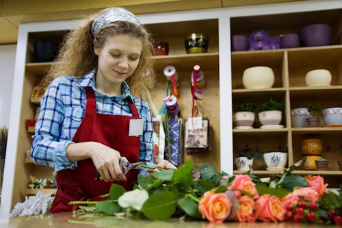 From below of positive female florist in apron and hairband using scissors for cutting low parts of rose branches placed on table in workspace against shelves with flower pots and decorations for presents