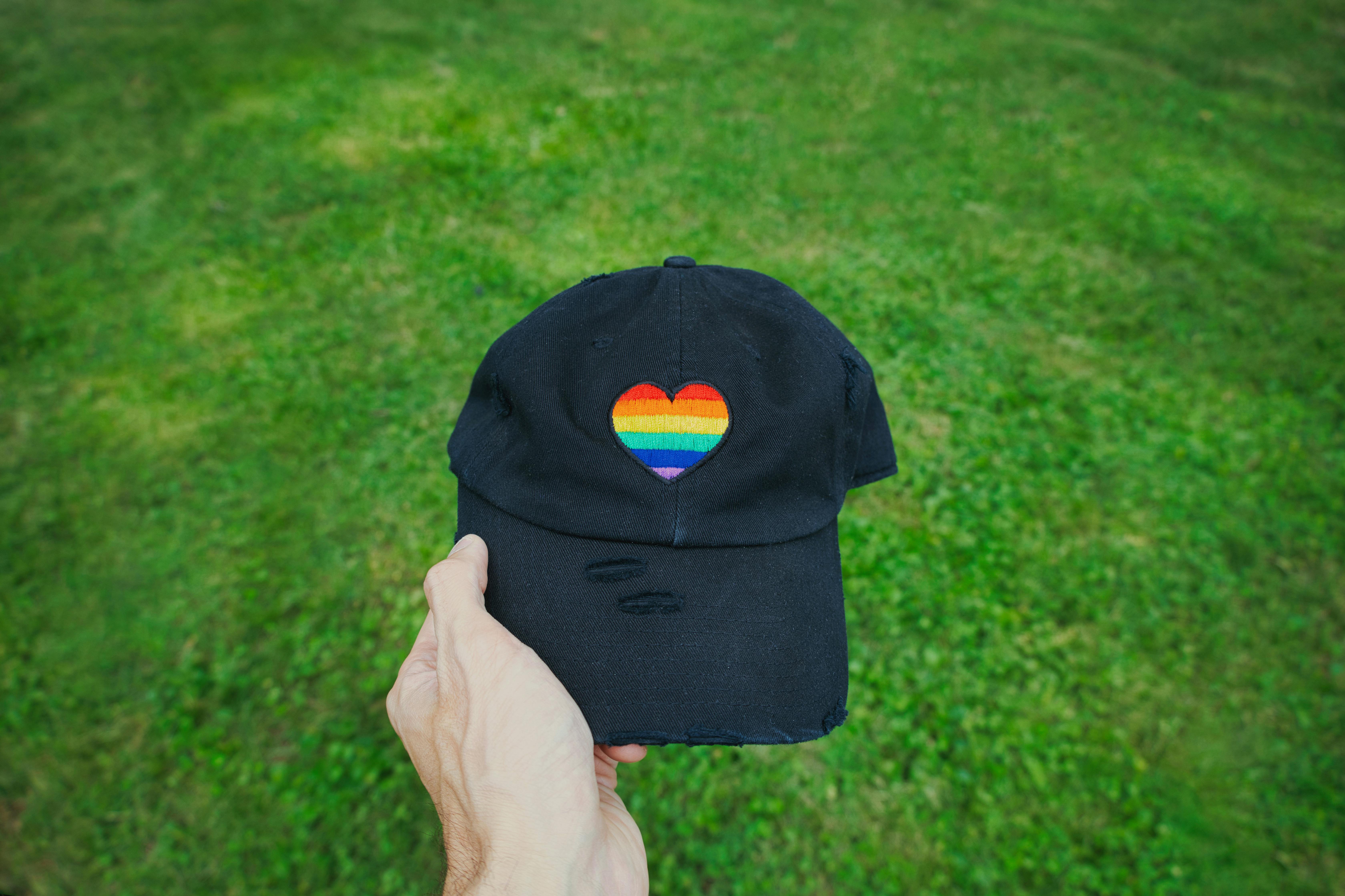 Hand holding a black cap with a rainbow heart, symbolic of pride, in a grassy field.