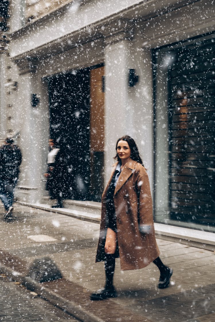 A Woman In Brown Trench Coat Walking On The Street Side During Snow Fall