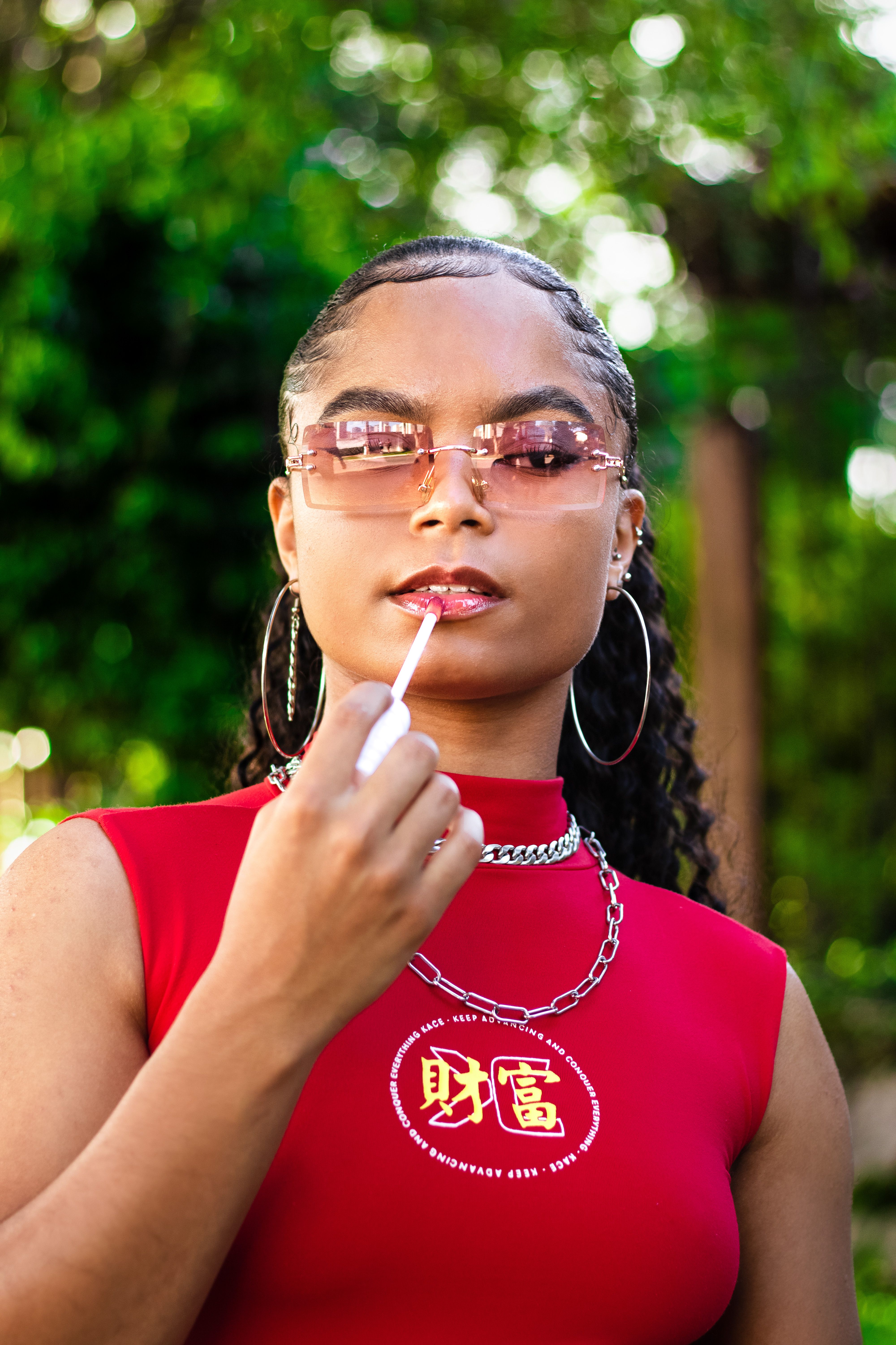 Confident woman in a red top applying lip gloss outdoors, wearing eyeglasses and jewelry.