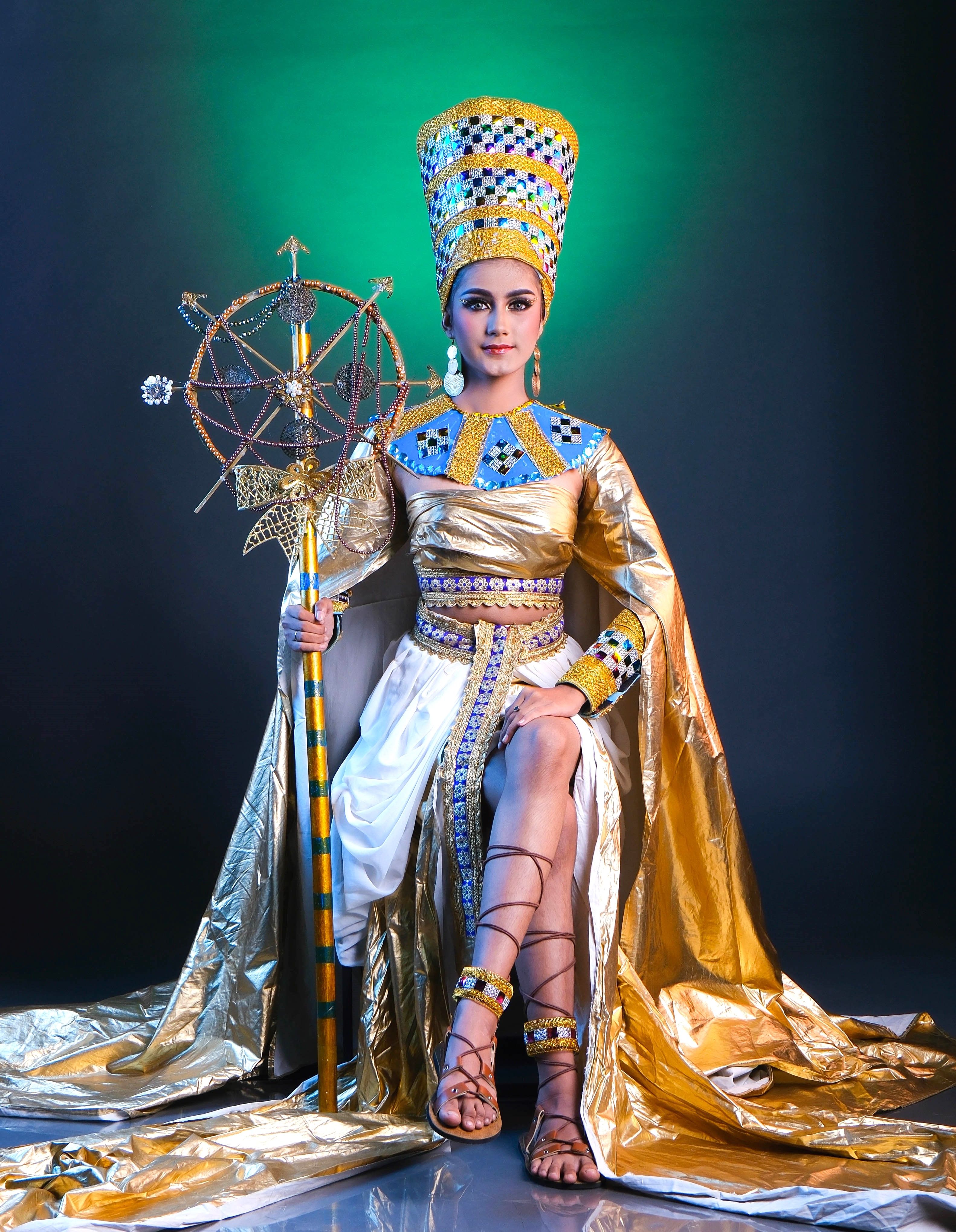 Elegant woman in gold traditional attire, sitting with a scepter against a green backdrop.