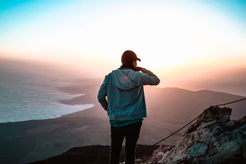 Free A man stands on a mountain peak, taking in the vibrant sunset view over the landscape. Stock Photo