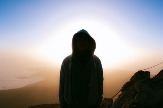 A dramatic silhouette of a person with a hoodie enveloped by a golden hour sunset atop a mountain.