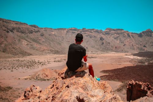 Free A man sitting on rocks observes a vast arid desert under a clear blue sky, captured from behind. Stock Photo