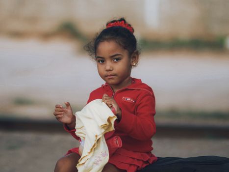 A young girl in a red top sitting outdoors holding a cloth, looking thoughtful.
