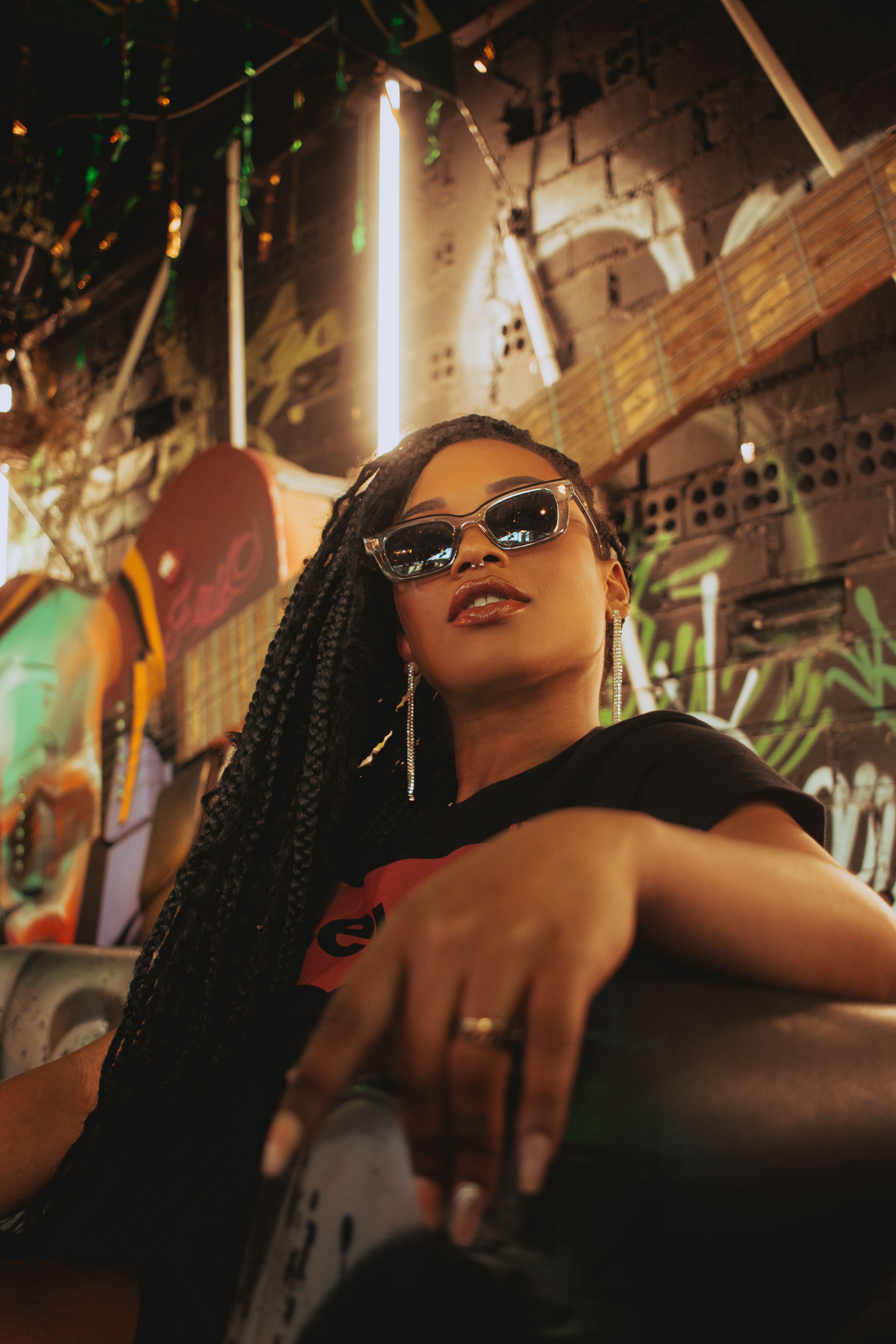 Fashionable woman with braided hair and sunglasses sitting indoors. Urban graffiti background.