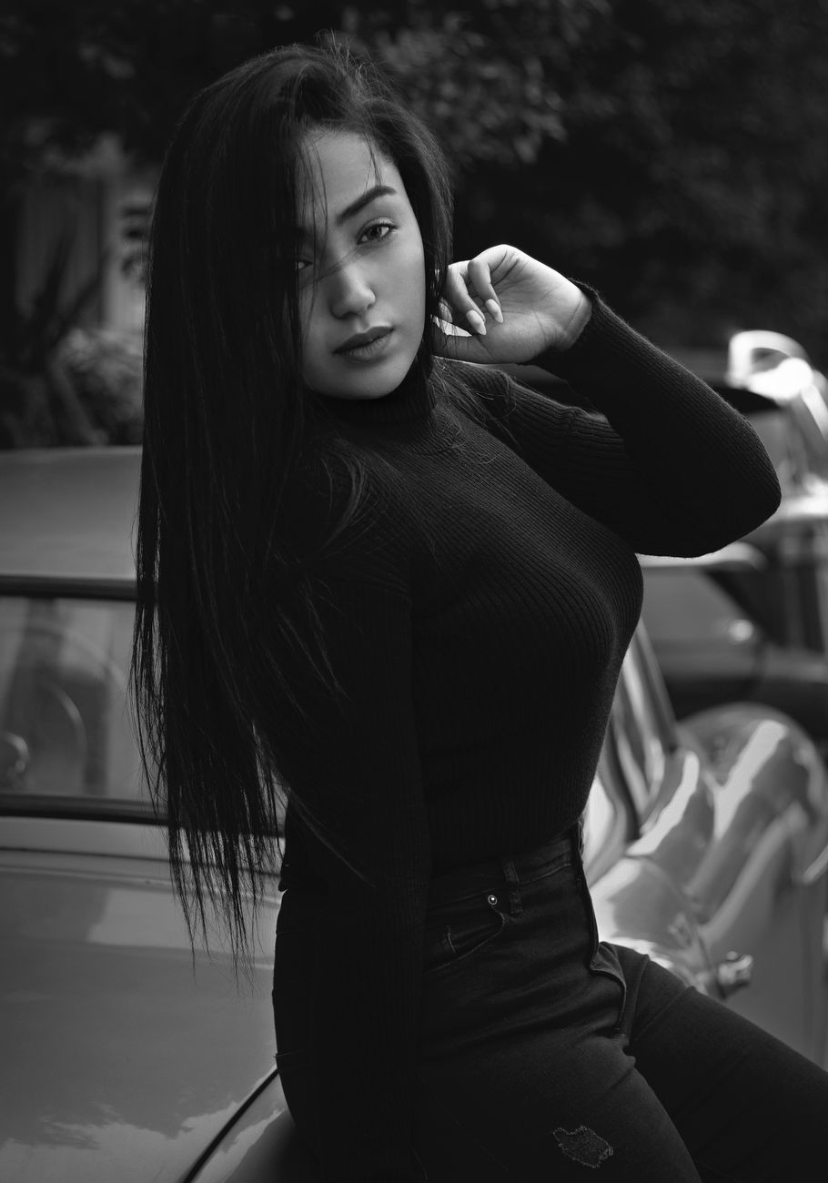 Black and white portrait of a fashionable young woman posing confidently by a vintage car.