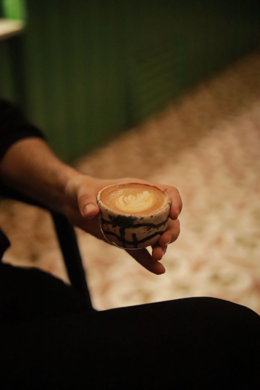 Close-up of a hand holding a cup of artisan latte with intricate latte art in a cozy indoor setting.. pexels, close, hand