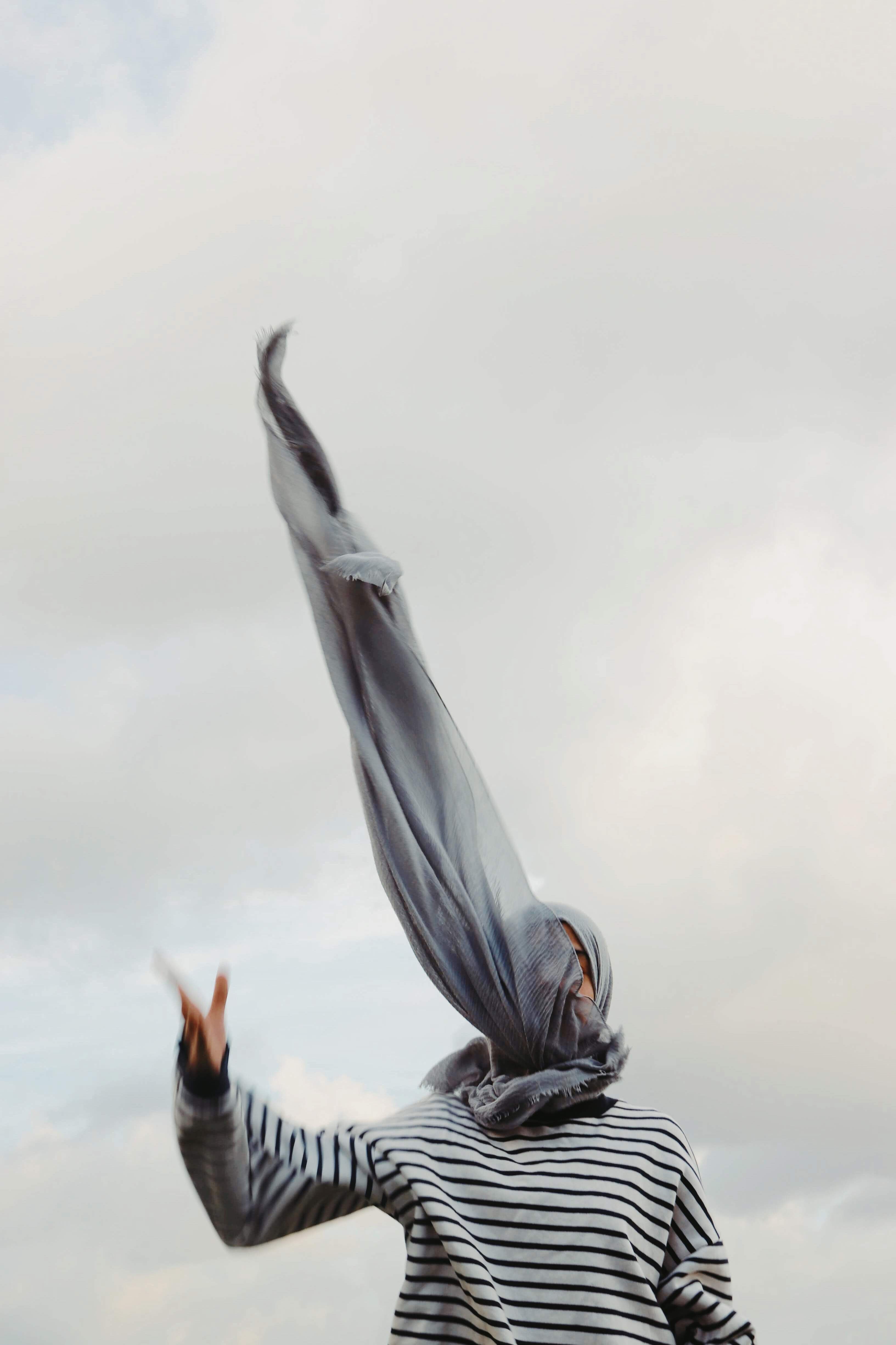Artistic shot of a woman outdoors with a flowing headscarf against a cloudy sky.