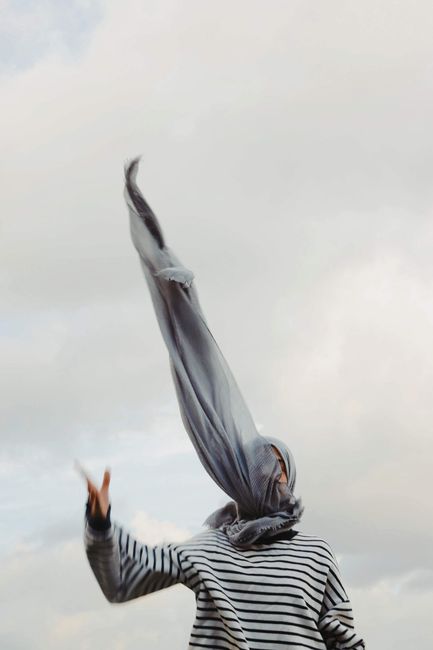 Artistic shot of a woman outdoors with a flowing headscarf against a cloudy sky.