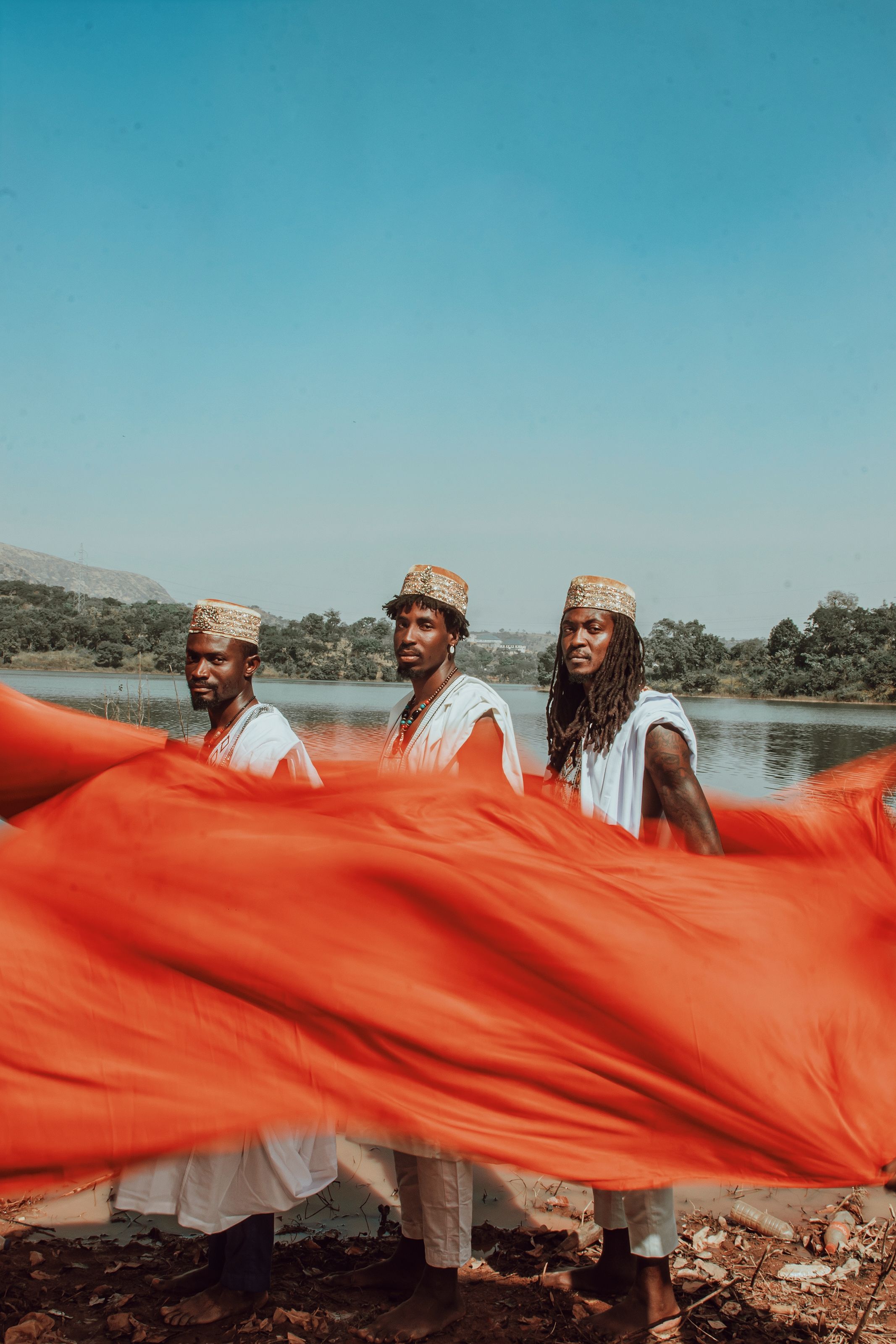 Three men posing by a river in traditional attire with a vibrant red fabric contrast