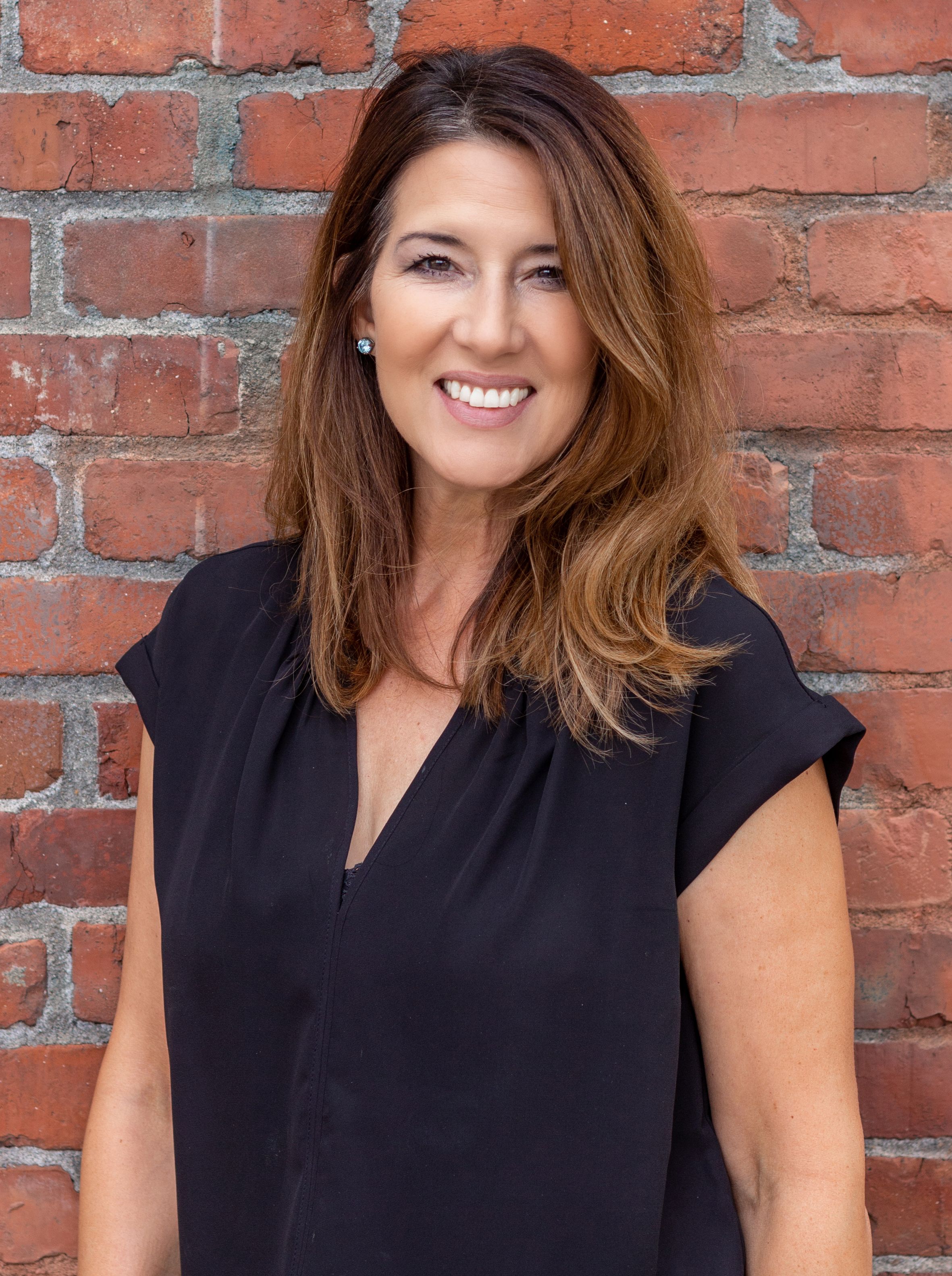 Portrait of a smiling woman in a black top standing in front of a brick wall.