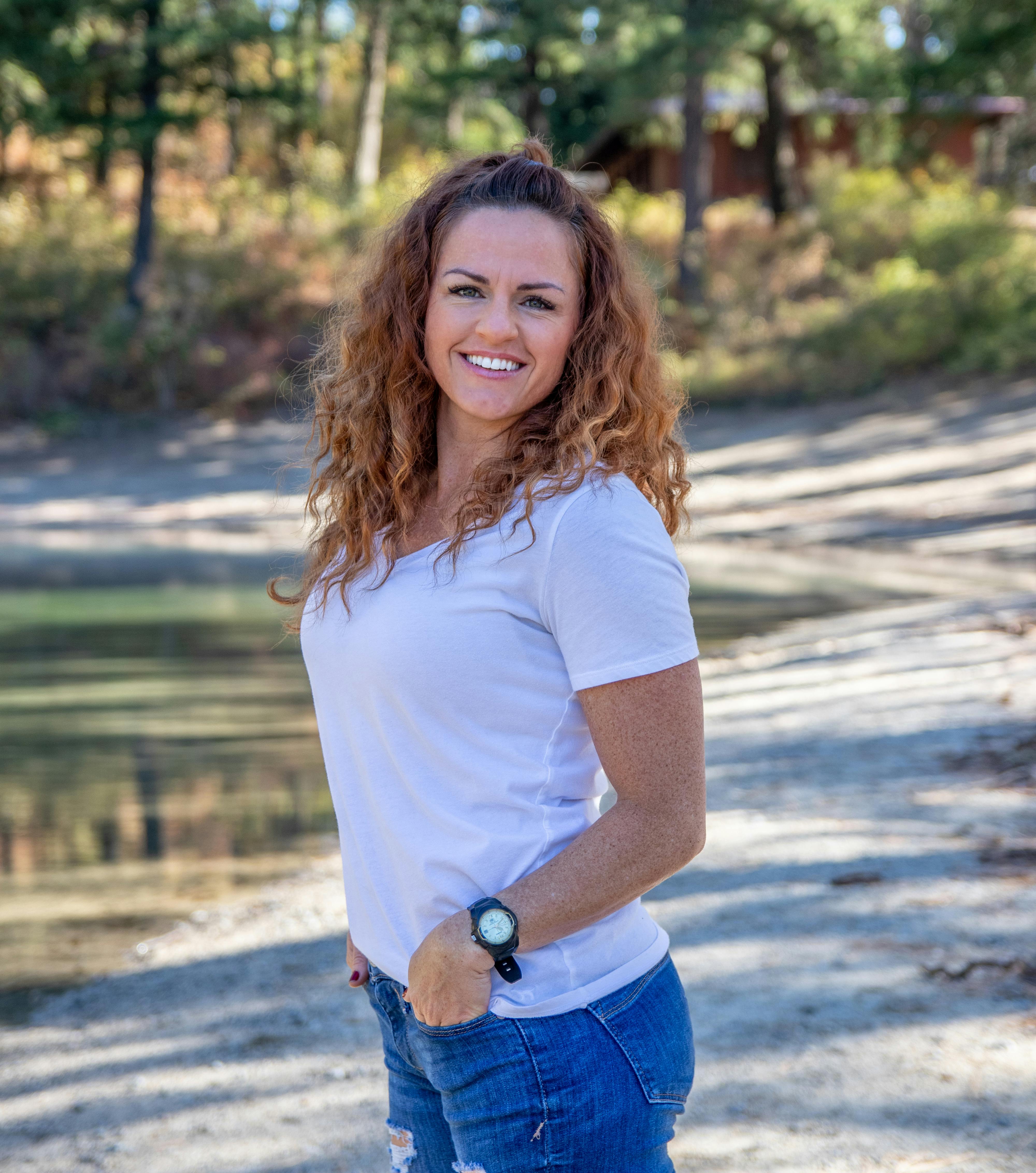 A cheerful woman in casual attire poses by a tranquil lake on a sunny day.