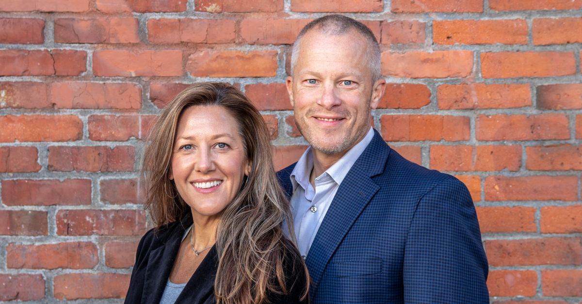 A cheerful middle-aged man and woman in business attire posing against a brick wall, depicting professionalism and partnership.