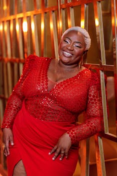 A stylish African American woman in a red dress posing confidently indoors with a vibrant smile.