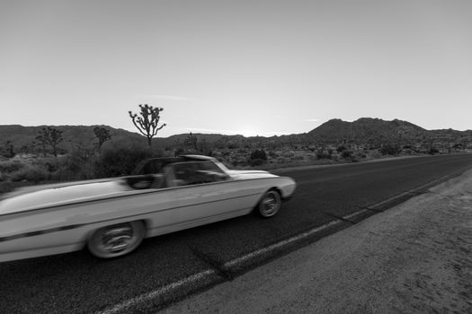 A classic convertible speeds through a desert road at twilight, showcasing motion in black and white.