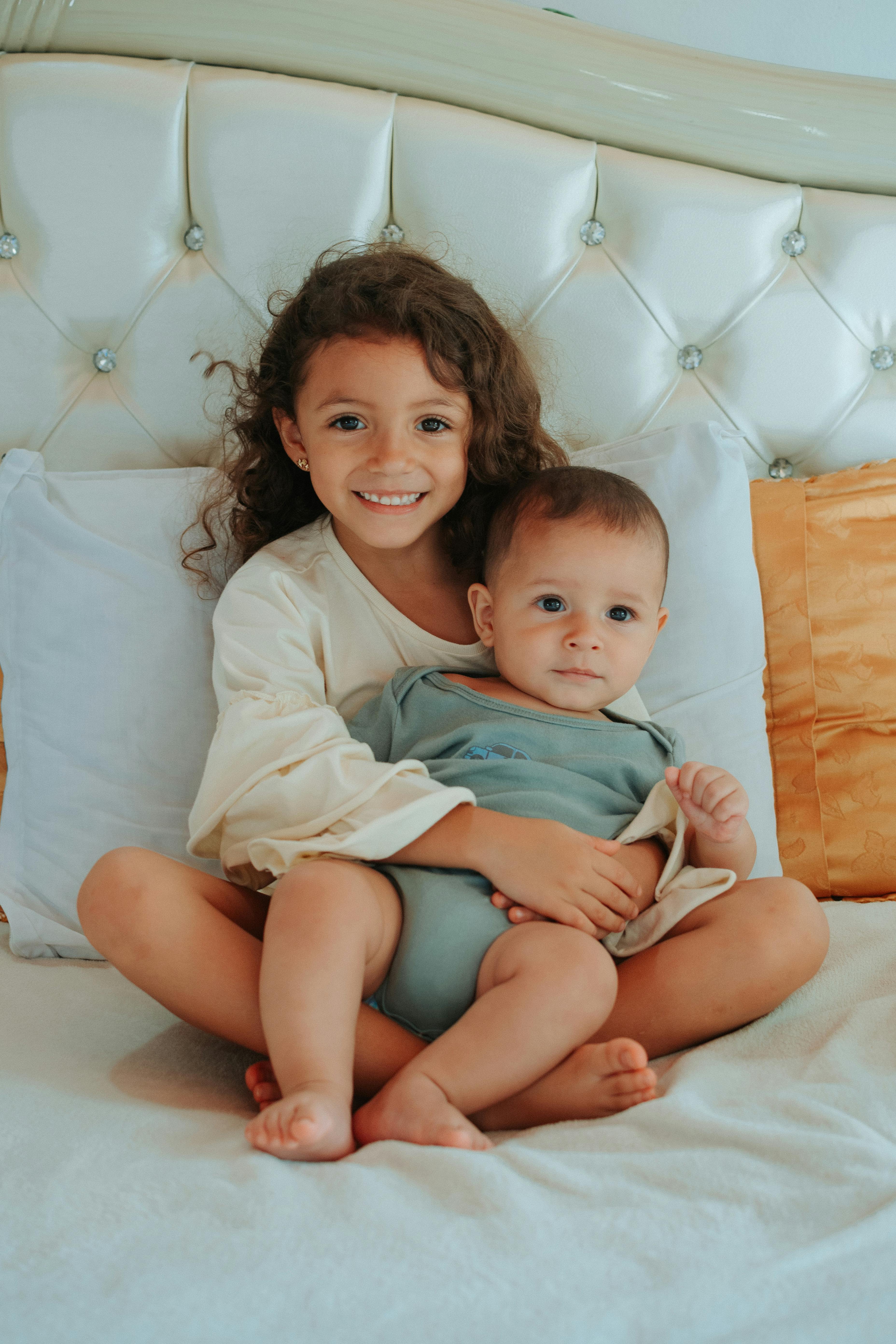A cheerful girl and her baby brother hugging while seated on a bed, capturing a moment of sibling love.