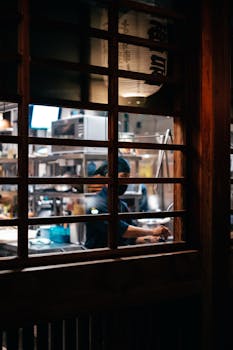 Atmospheric view of a chef working in a dimly lit restaurant kitchen through a wooden window frame.