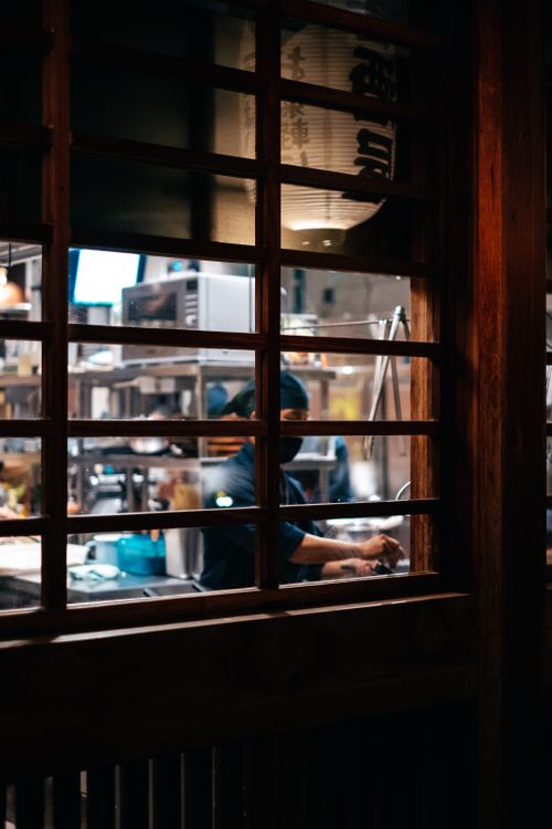 Free Atmospheric view of a chef working in a dimly lit restaurant kitchen through a wooden window frame. Stock Photo