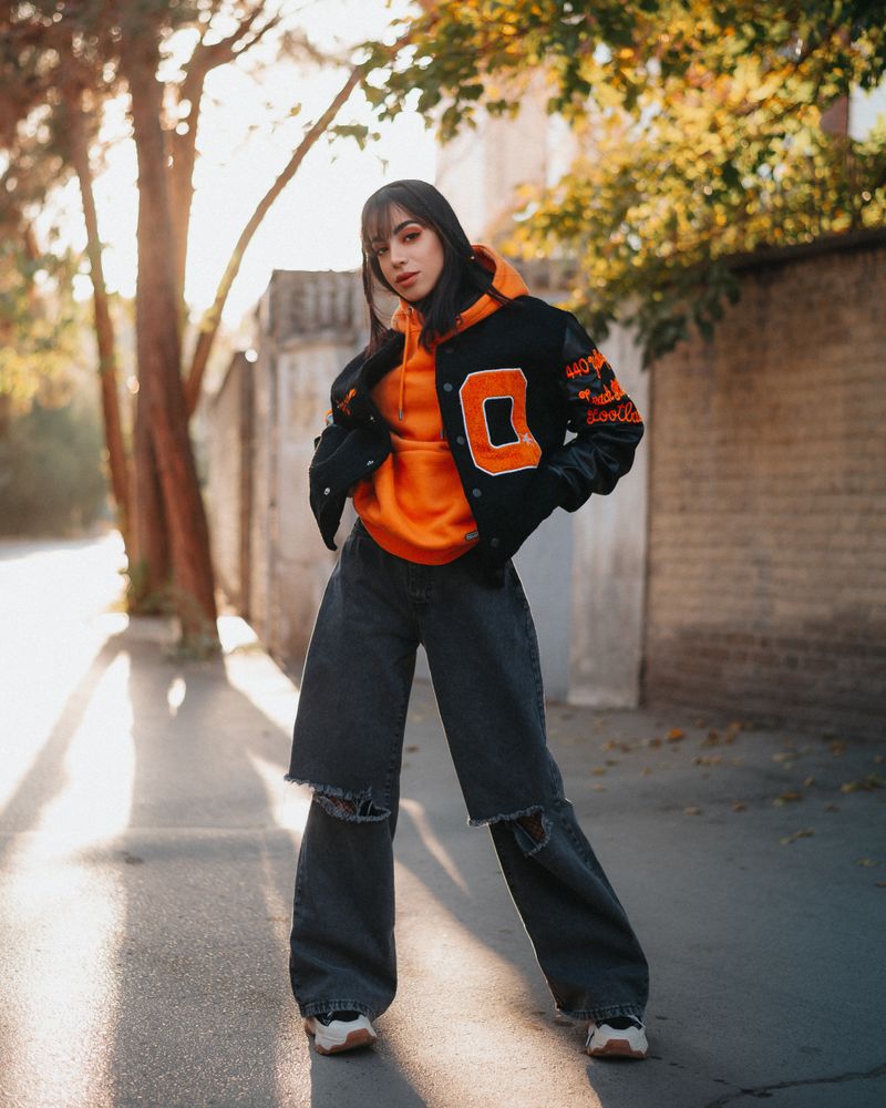 Woman in orange hoodie, varsity jacket and ripped jeans