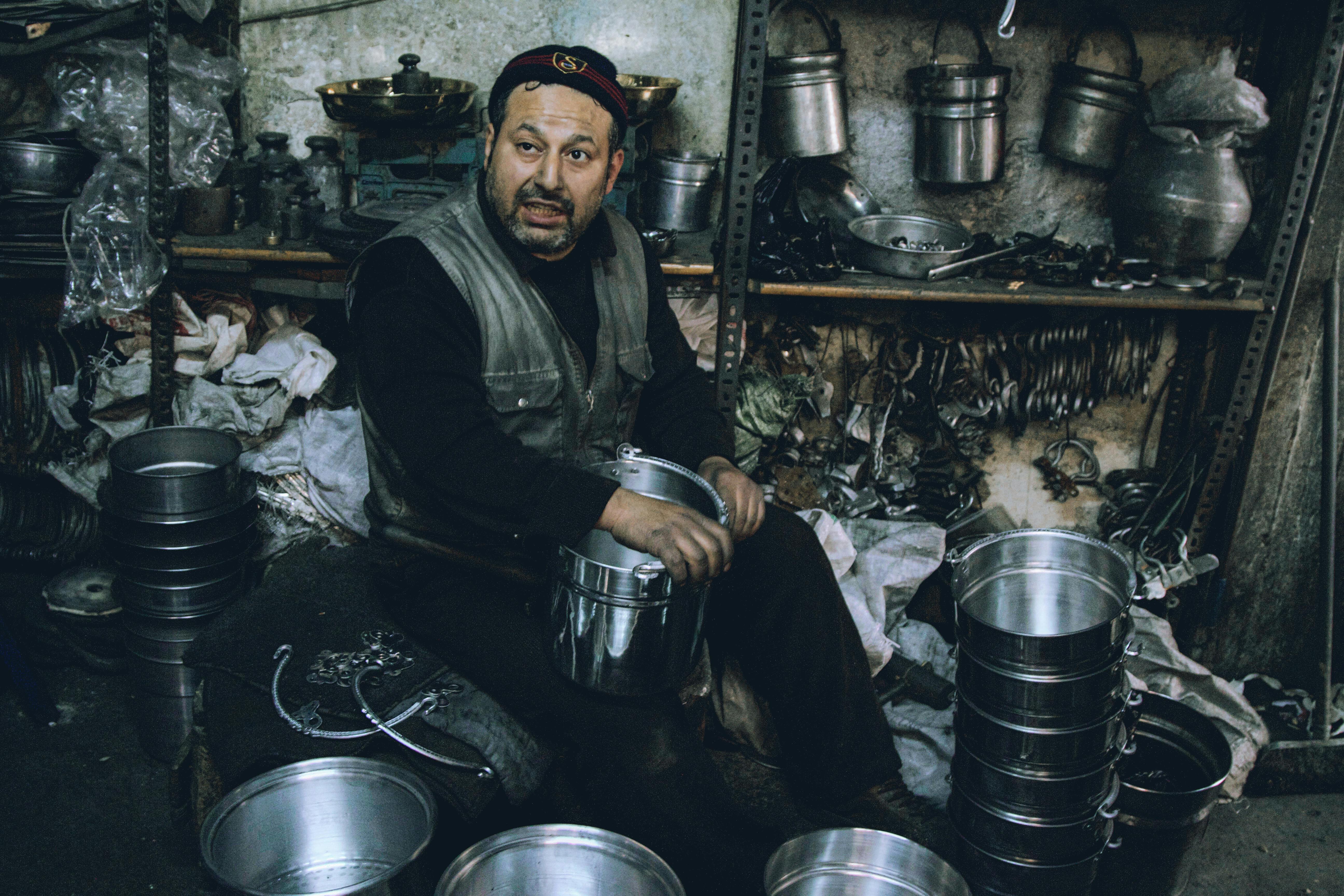 A craftsman sitting in a metal workshop with pots and tools displaying traditional craftsmanship.